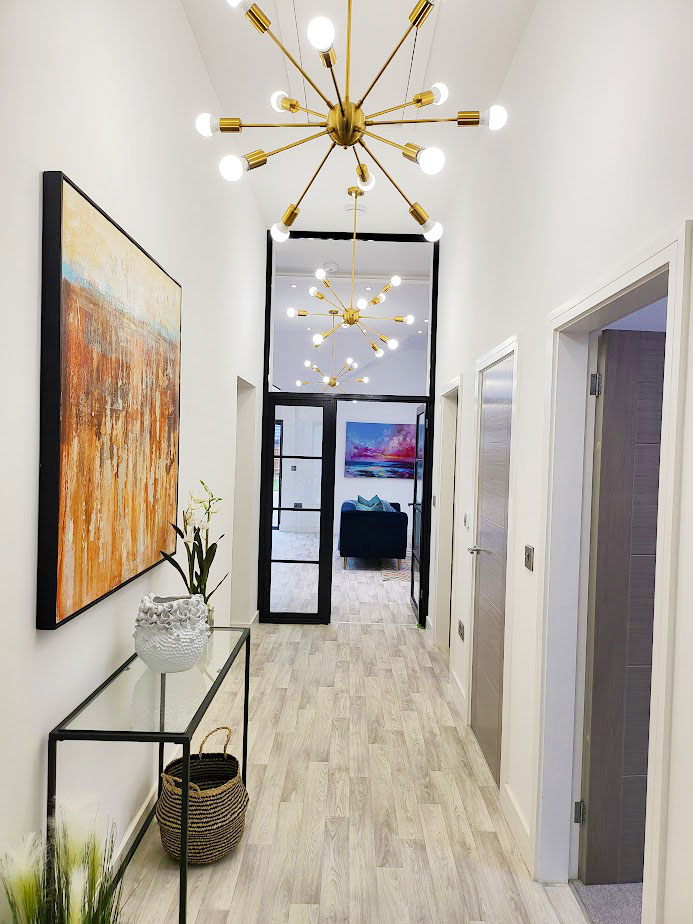 Home Staging For Sale Example. Hallway with new grey flooring, bold gold chandelier lighting, black and glass console table on the left with large orange and pale blue art work above.